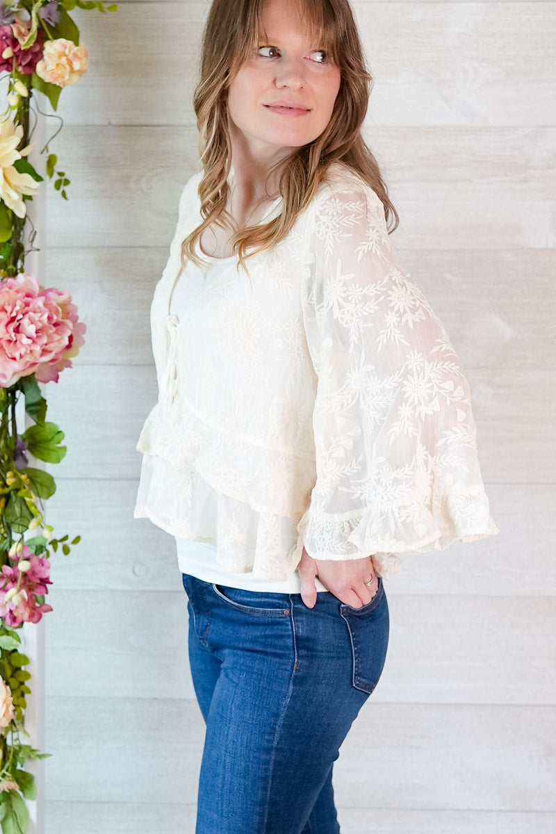 Woman wearing a white lace blouse and blue jeans standing next to floral decorations.