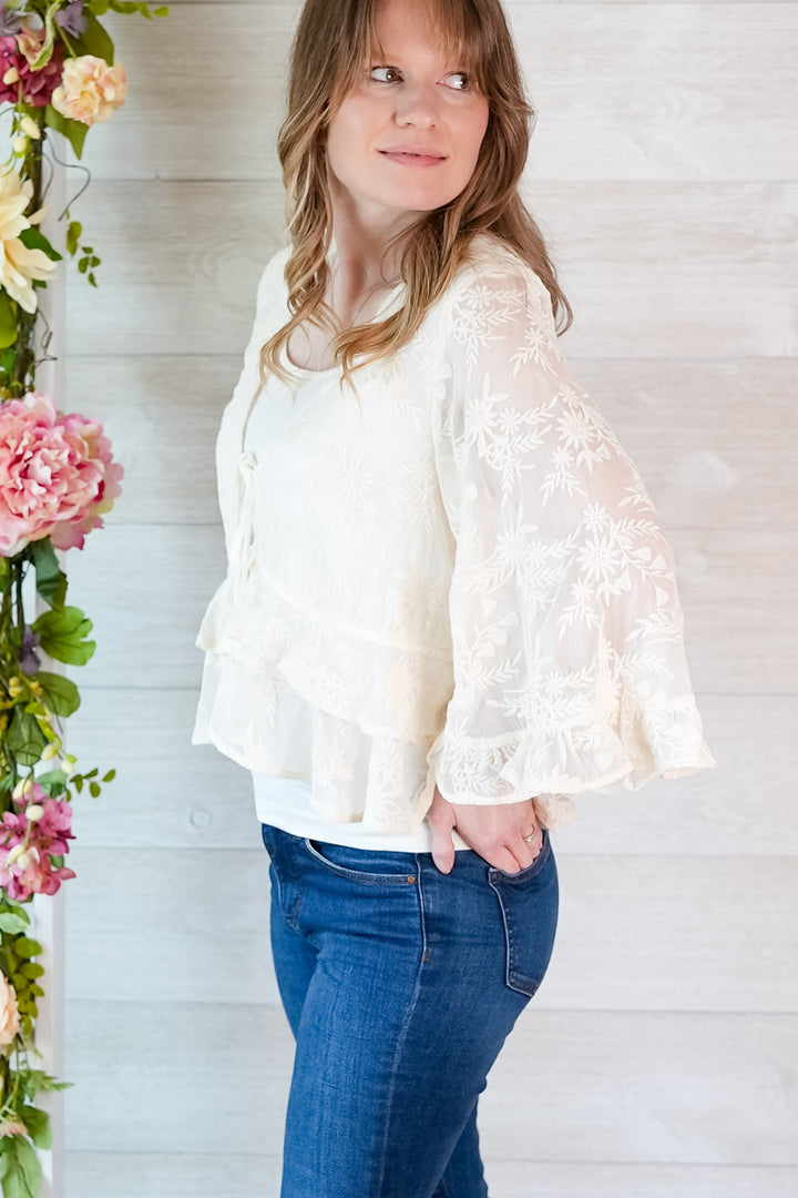 Woman wearing a white lace blouse and blue jeans standing next to floral decorations.