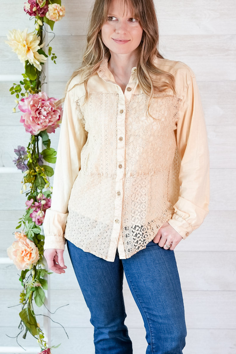 Woman wearing a light-colored blouse with lace details and blue jeans, standing next to floral decorations against a white wooden background.