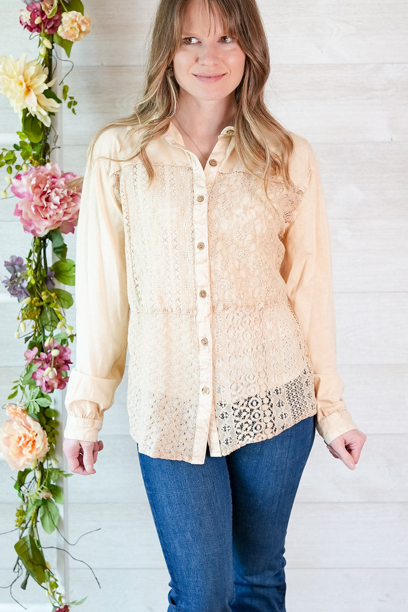 Woman wearing a beige lace-trimmed blouse with blue jeans, standing next to floral decorations.