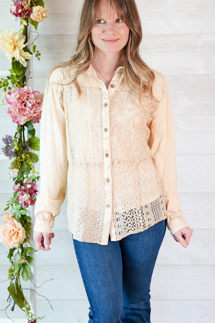 Woman wearing a beige lace-trimmed blouse with blue jeans, standing next to floral decorations.