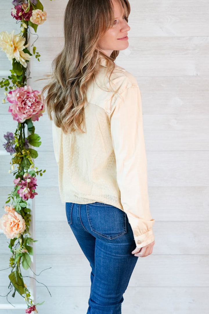 Woman wearing a light-colored blouse and blue jeans standing next to a floral arrangement against a white wooden panel background.