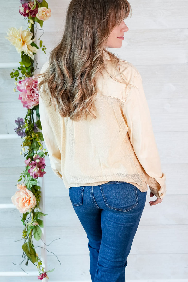 Woman wearing a light-colored blouse and blue jeans standing next to floral decorations against a white wall.