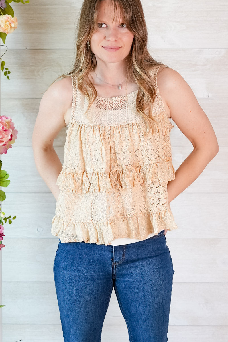 Woman wearing a beige tiered lace tank top and blue jeans against a white wooden background.
