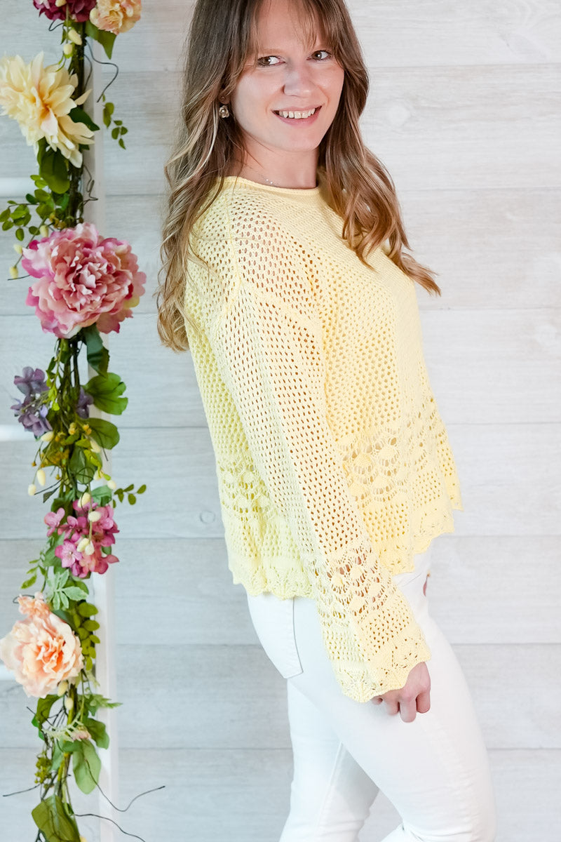Woman wearing a yellow crochet top standing next to floral decorations against a white wooden wall.