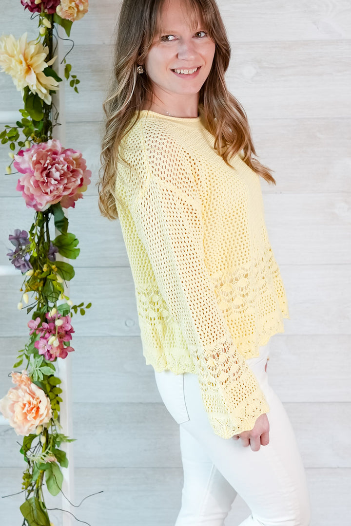 Woman wearing a yellow crochet top standing next to floral decorations against a white wooden wall.