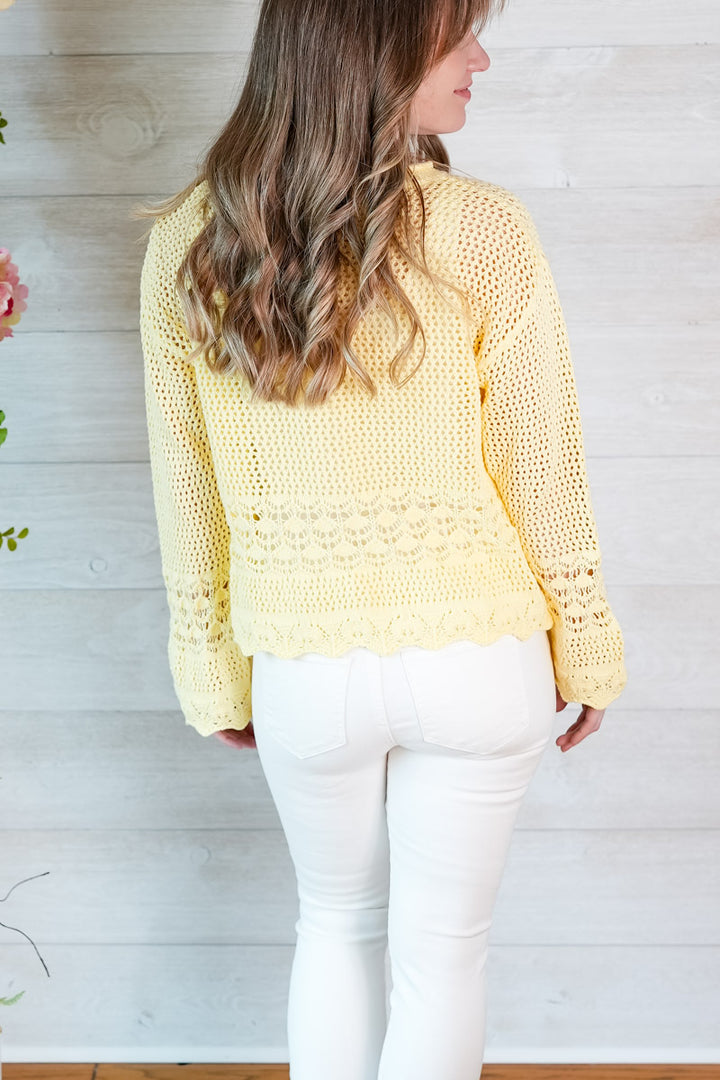 Woman wearing a yellow crochet sweater and white pants against a wooden background