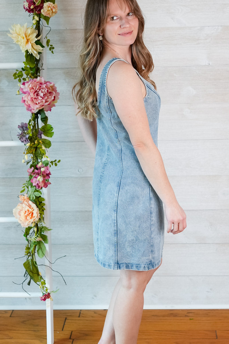 Woman wearing a light wash denim mini dress standing next to a floral arrangement against a white wall.