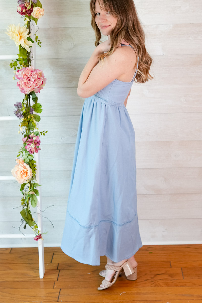 Woman in a light blue dress standing next to a floral arrangement against a wooden floor and white wall.