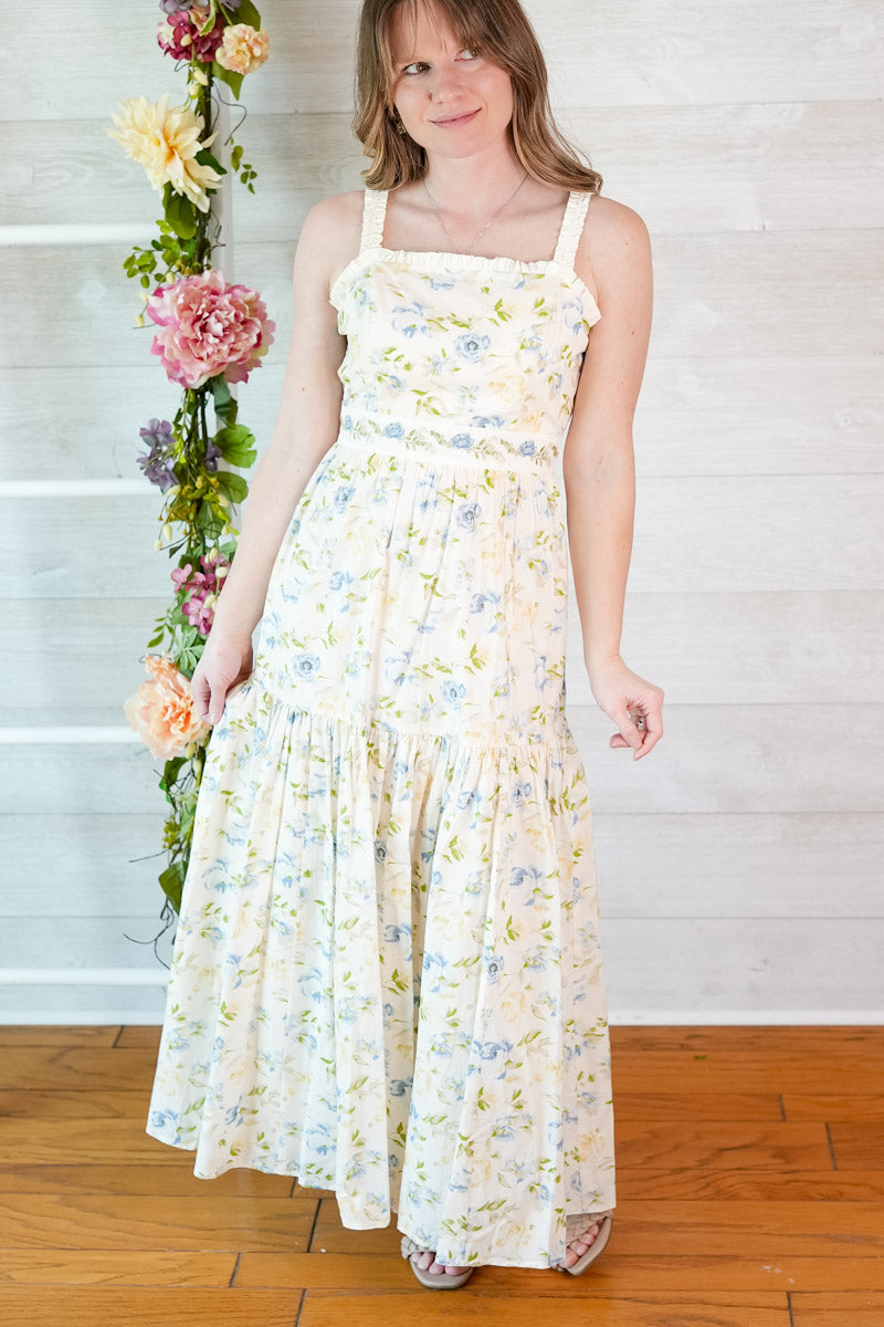 Woman wearing a blue and yellow tiered floral midi dress standing next to a floral arrangement indoors.