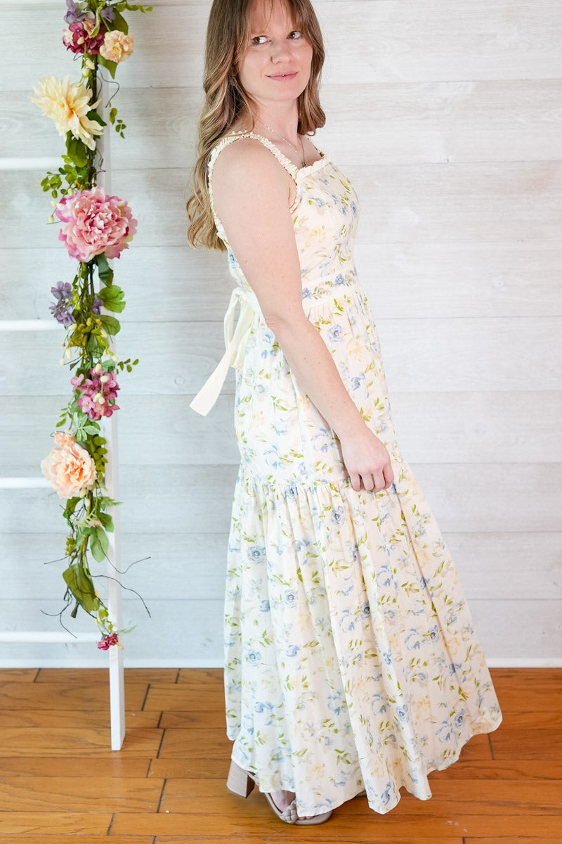 Woman in a blue and yellow floral dress standing next to a decorative floral arrangement against a white wooden panel background.
