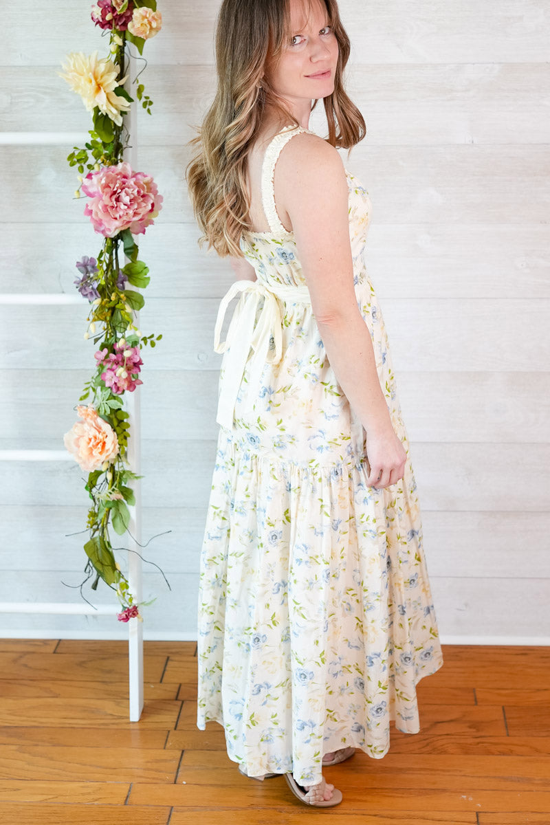 Woman in a blue and yellow spring floral dress standing next to a floral arrangement against a white wall.