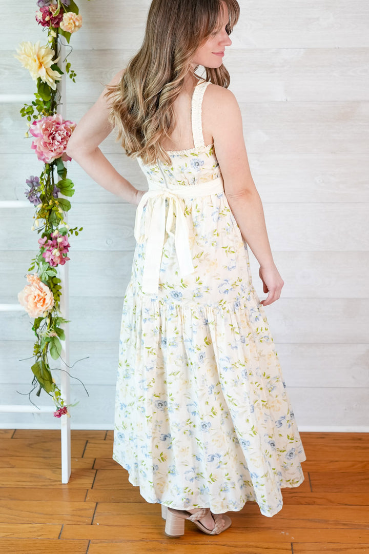 Woman wearing a blue and yellow spring floral dress with back bow tie standing next to a white ladder with flowers against a white wall.