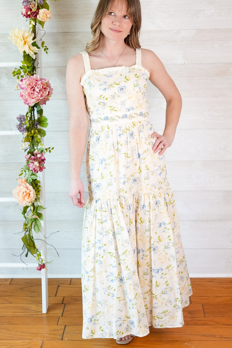 Woman wearing a yellow and blue tiered floral midi dress standing next to a floral arrangement indoors.