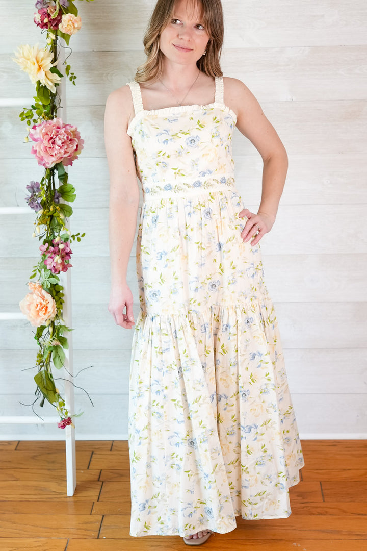 Woman wearing a yellow and blue tiered floral midi dress standing next to a floral arrangement indoors.