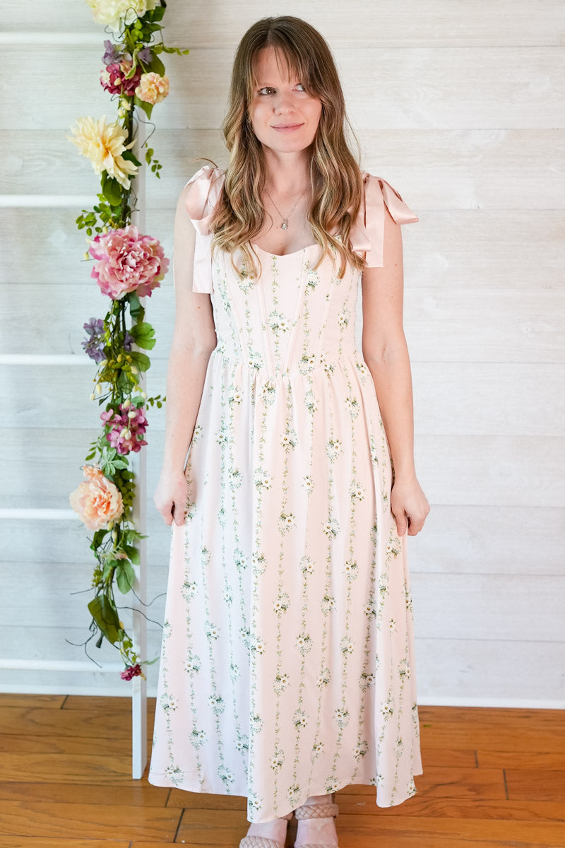 Woman wearing a pink floral striped midi dress with ribbon straps standing in front of a white wall with flowers.