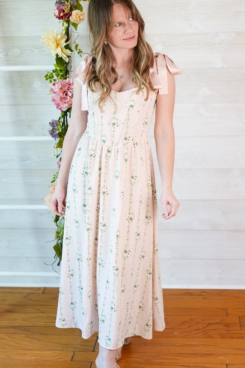 Woman wearing a pink floral striped midi dress with ribbon straps standing in front of a white wall with flowers.