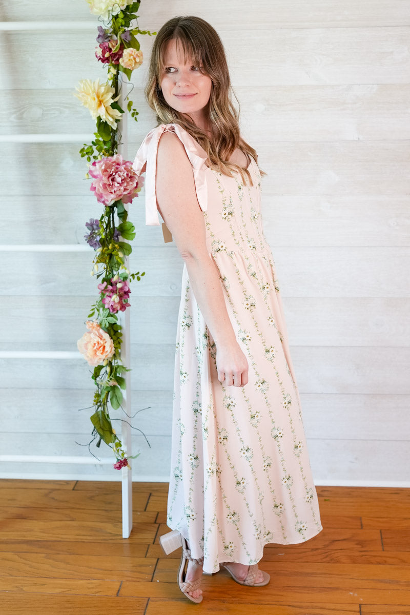 Woman wearing a pink floral striped midi dress with ribbon straps standing in front of a white wall with flowers.