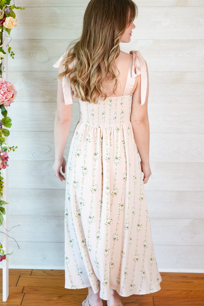 Woman wearing a pink floral striped midi dress with ribbon straps standing in front of a white wall with flowers.