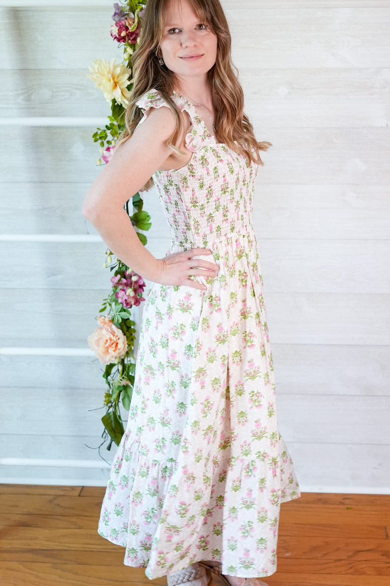 Woman wearing a green and pink floral tiled midi dress standing next to flowers against a white wooden panel background