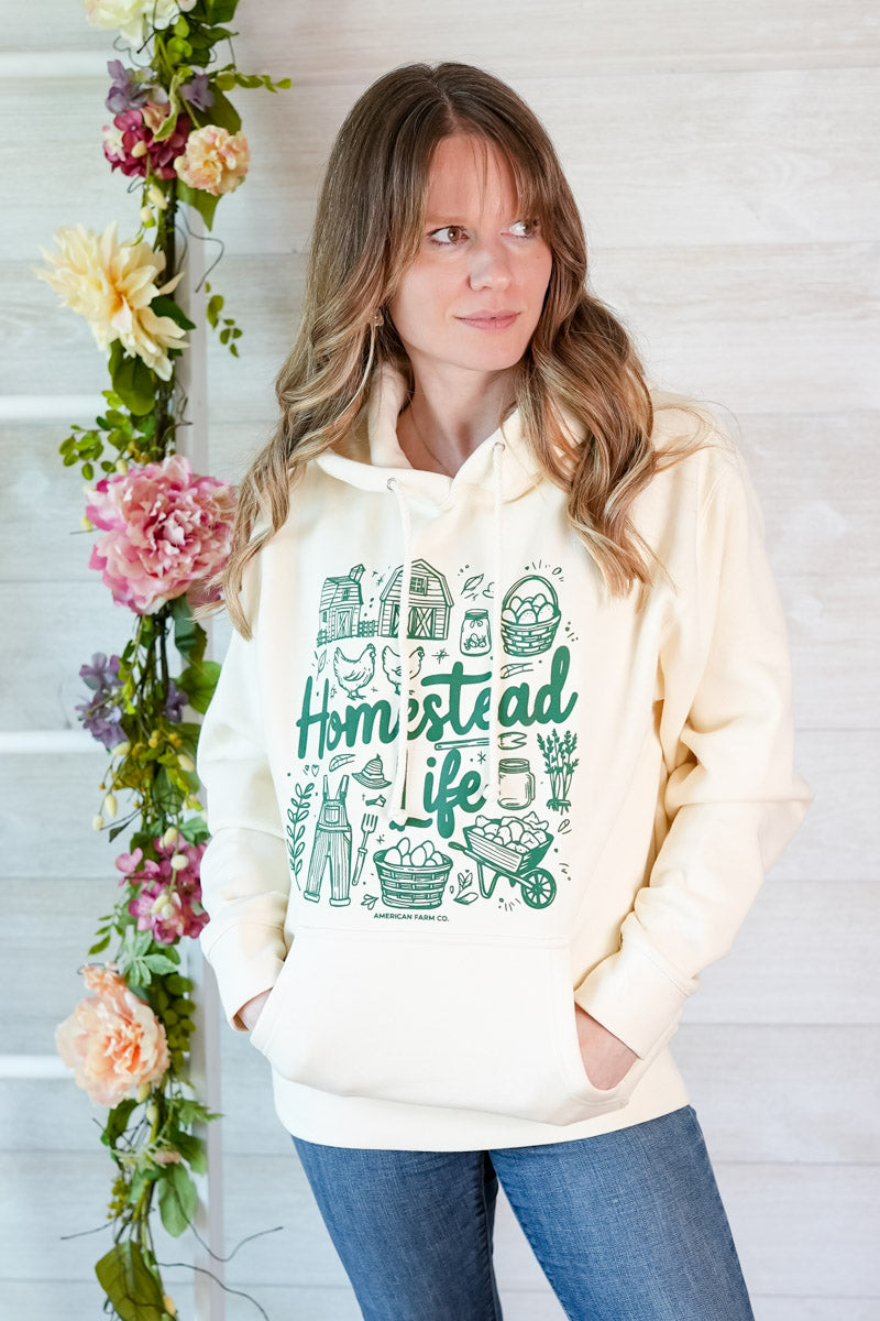 Woman wearing a cream hoodie that says Homestead Life and has green country inspired graphic icons