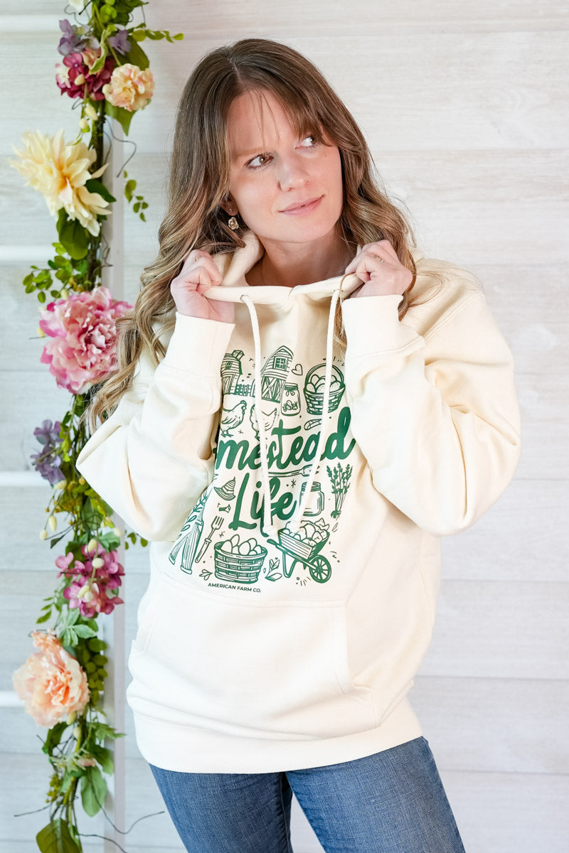 Woman wearing a hoodie with 'Homestead Life' text and graphics, standing next to floral decorations.