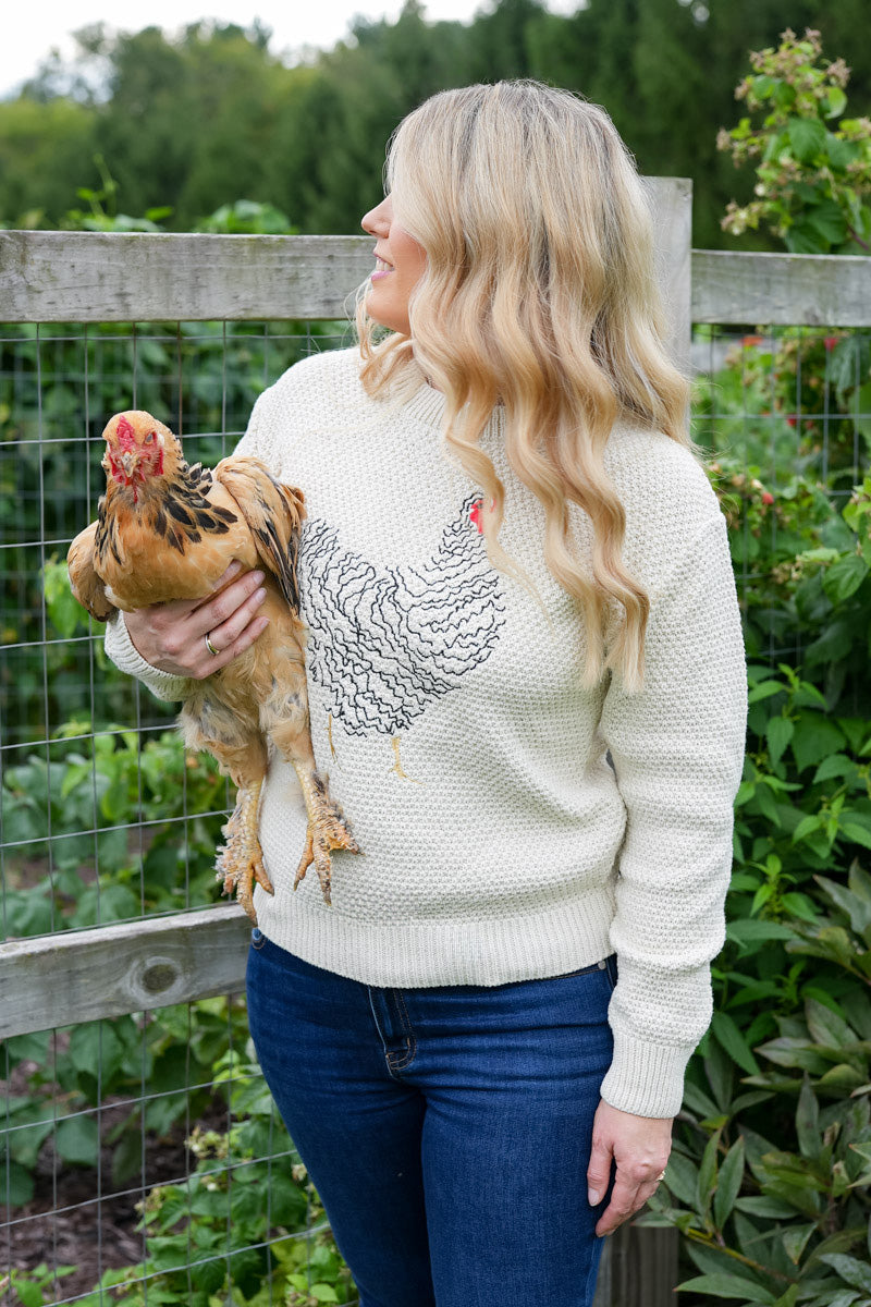 A woman standing in a garden holding a brown chicken, wearing a cream knit sweater with a black rooster graphic and blue jeans.