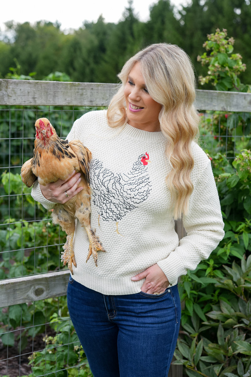 A woman standing outdoors in a garden holding a brown chicken, wearing a cream-colored knit sweater with a black and red chicken graphic on the front, paired with blue jeans.