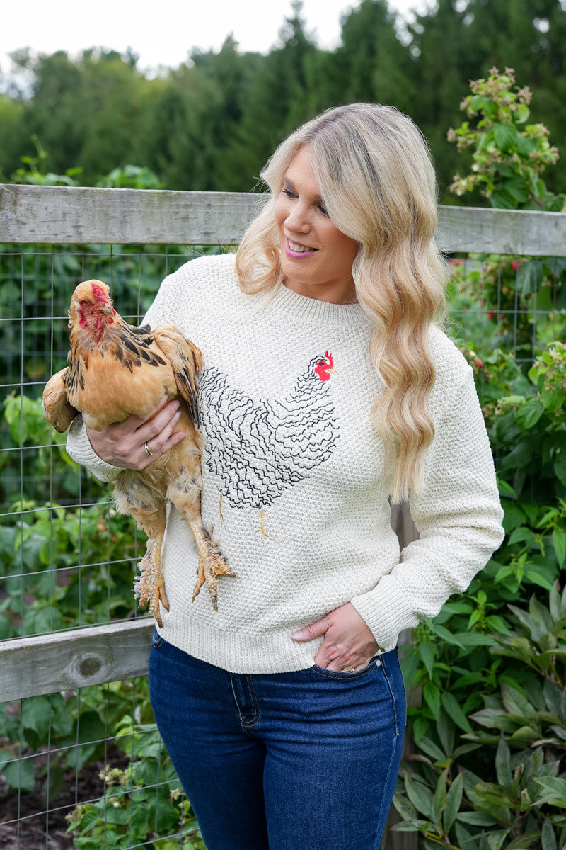 A woman standing in a garden holding a brown chicken, wearing a cream-colored waffle-knit sweater with a black and white rooster graphic on the front, paired with blue jeans.