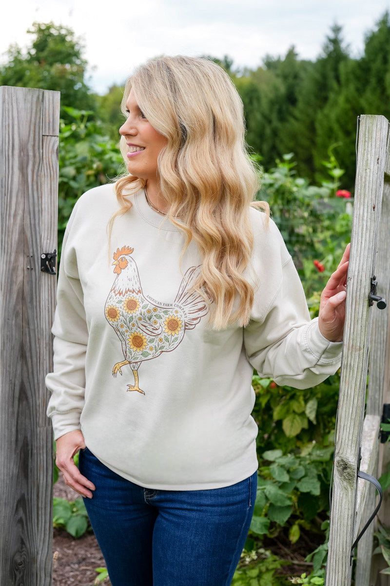 A woman with long blonde hair stands in a garden wearing a cream-colored crewneck sweatshirt with a colorful embroidered rooster and sunflower design, paired with blue jeans.