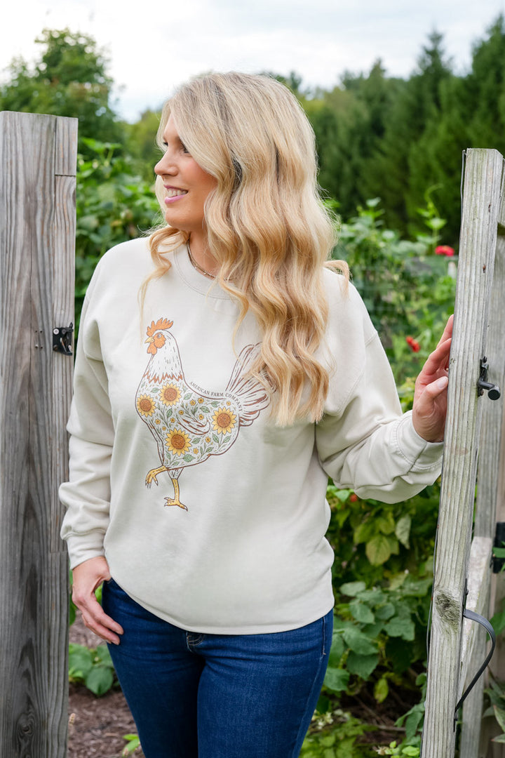 A woman with long blonde hair stands in a garden wearing a cream-colored crewneck sweatshirt with a colorful embroidered rooster and sunflower design, paired with blue jeans.