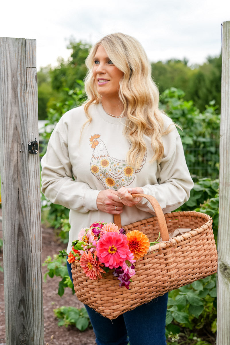 A woman with long blonde hair stands in a garden holding a wicker basket filled with colorful flowers. She is wearing a cream sweatshirt with a chicken and sunflower graphic, blue jeans, and is framed by wooden posts.