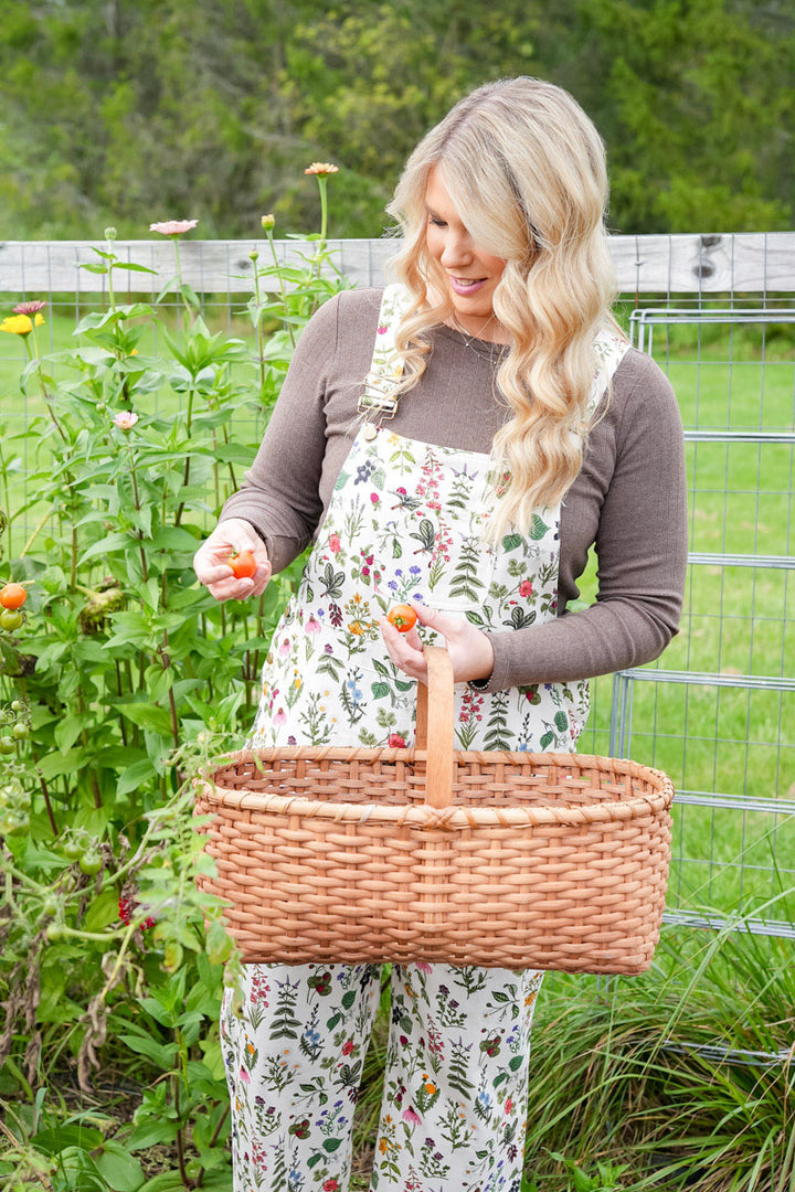 A woman standing in a garden holding a large woven wicker basket, wearing a brown long-sleeve shirt and white overalls with a colorful floral and butterfly print.