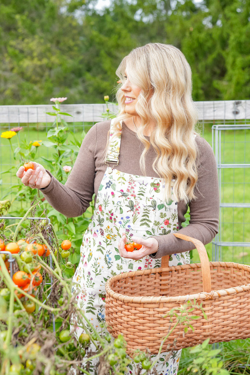 A woman with long blonde hair stands in a garden holding a wicker basket and picking tomatoes. She is wearing a brown long-sleeve shirt and a cream apron with a colorful floral and botanical print.