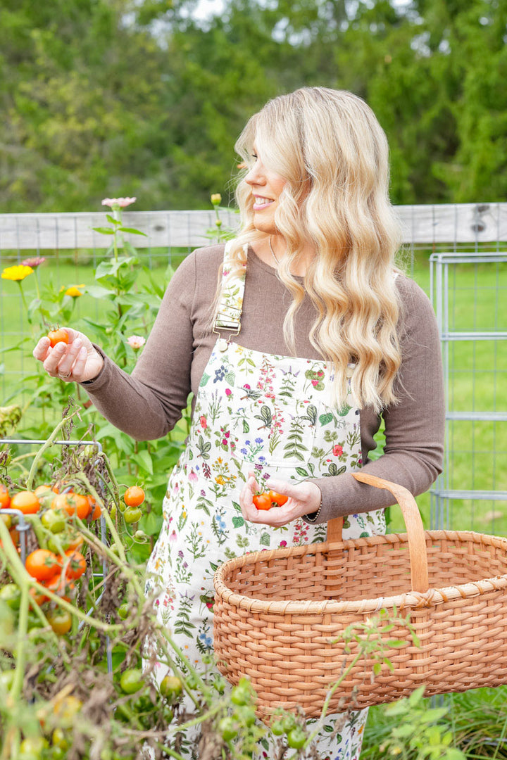 A woman with long blonde hair stands in a garden holding a wicker basket and picking tomatoes. She is wearing a brown long-sleeve shirt and a cream apron with a colorful floral and botanical print.