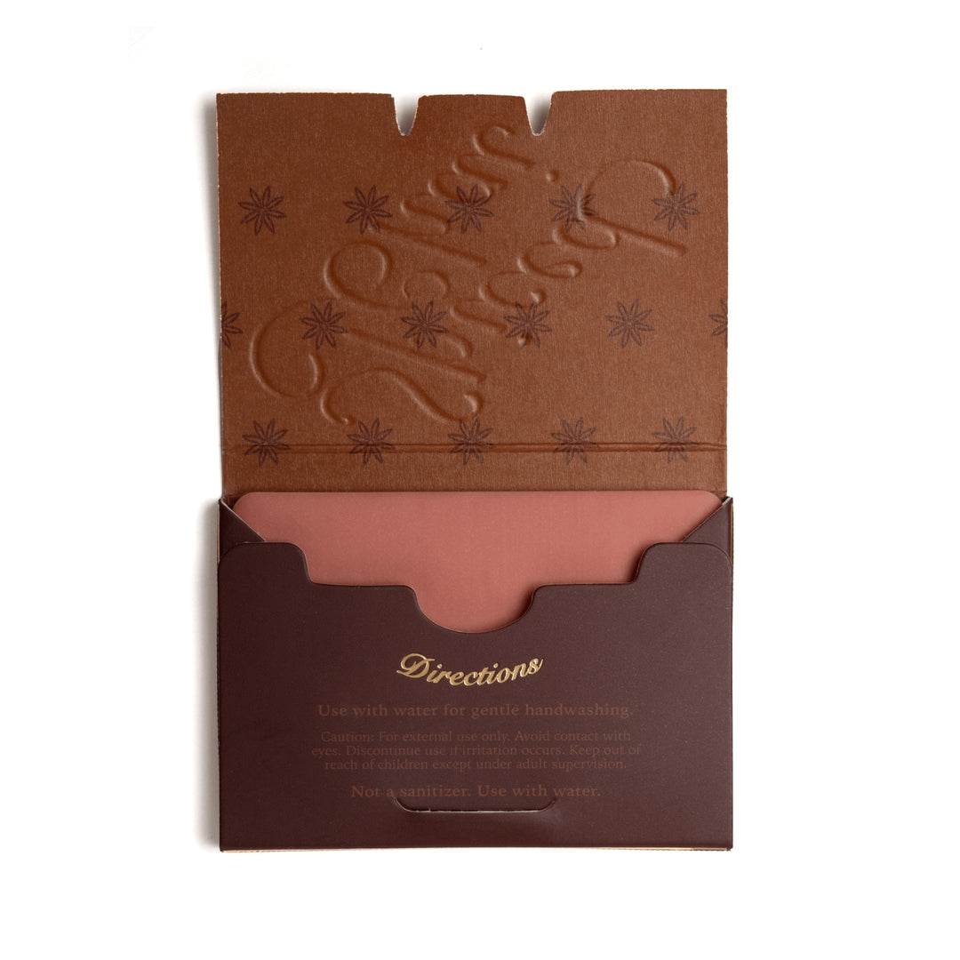 A brown kraft paper hand wash pouch with a matching embossed lid, shown open on a white background. The inside of the lid features an embossed design, and the bottom compartment contains a dusty rose-colored hand wash powder with printed usage instructions.