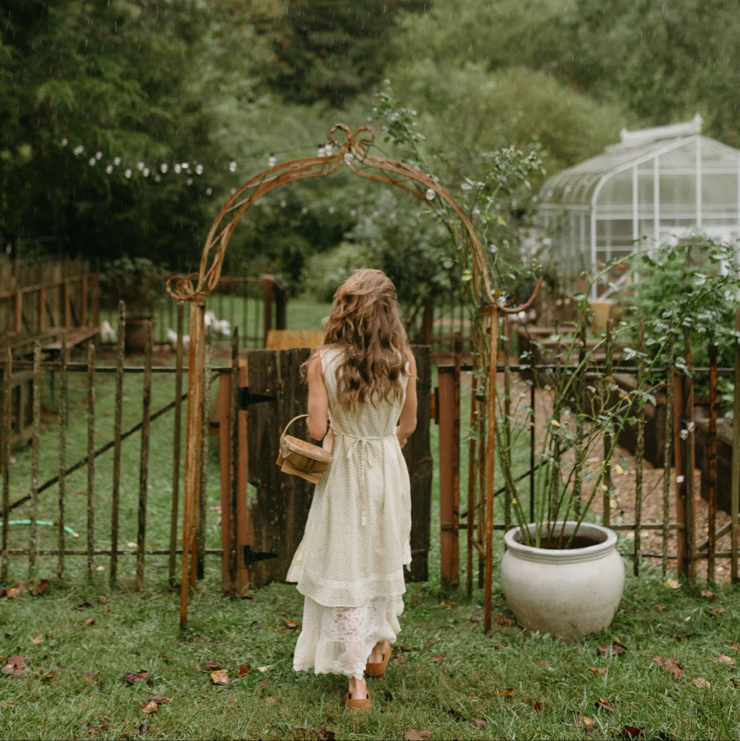 Woman in a white dress walking towards a rustic archway in a garden setting.