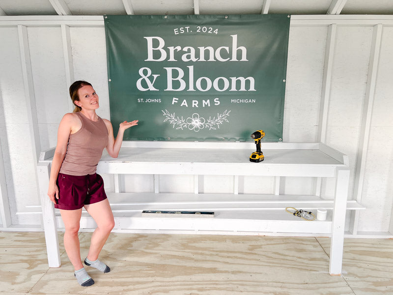 Person standing next to a white bench with a green 'Branch & Bloom' banner in a shed.