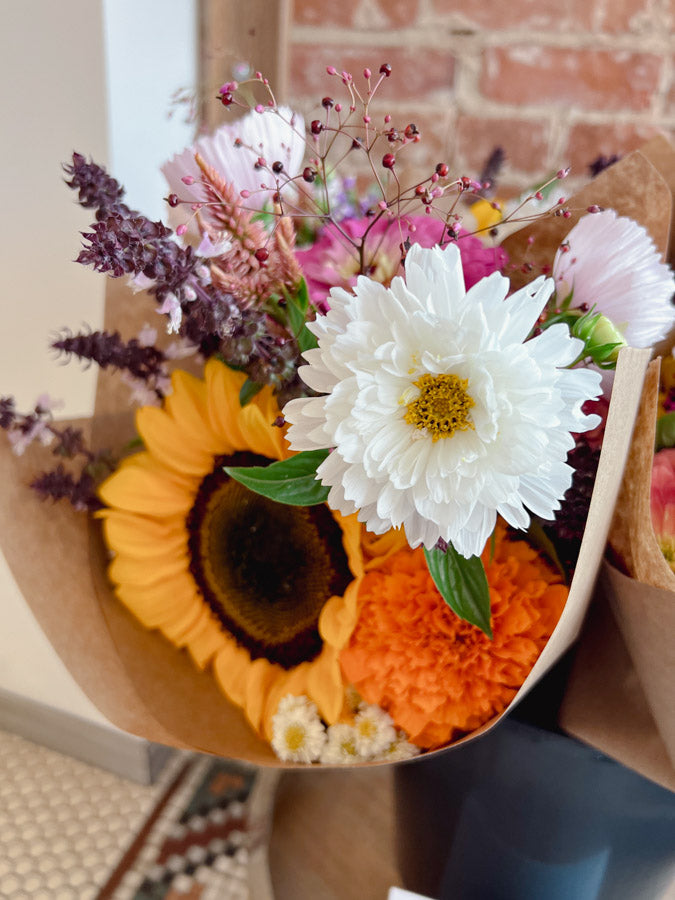 Bouquet of flowers with sunflowers, daisies, and other varieties against a brick wall.