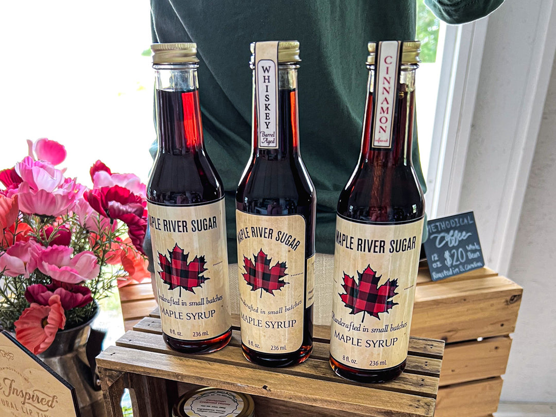 Three bottles of Maple River Sugar maple syrup on a wooden crate with flowers in the background.