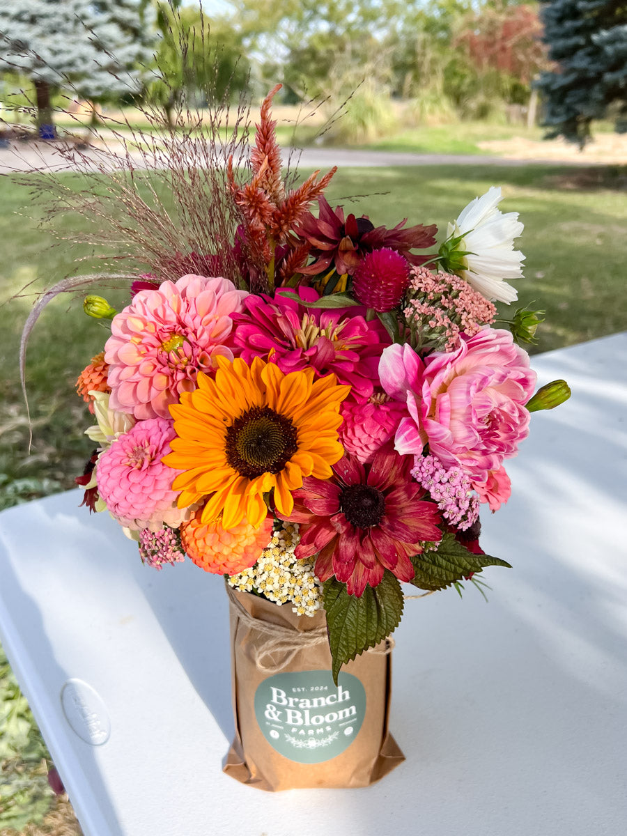 Bouquet of flowers in a wrapped mason jar with 'Branch & Bloom Farms' branding on a white table outdoors.