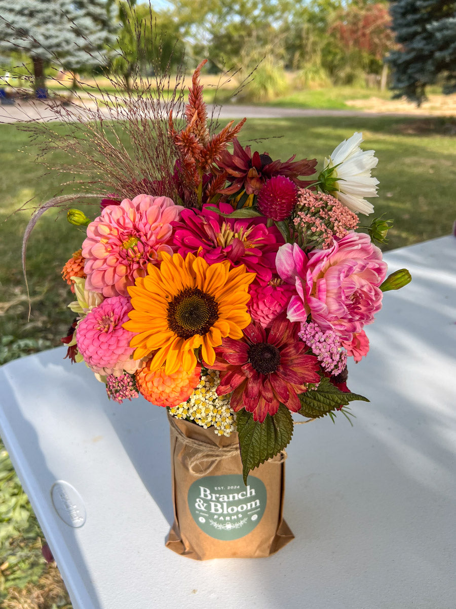 Colorful wrapped Mason jar flower bouquet with 'Branch & Bloom Farms' label on a white table outdoors