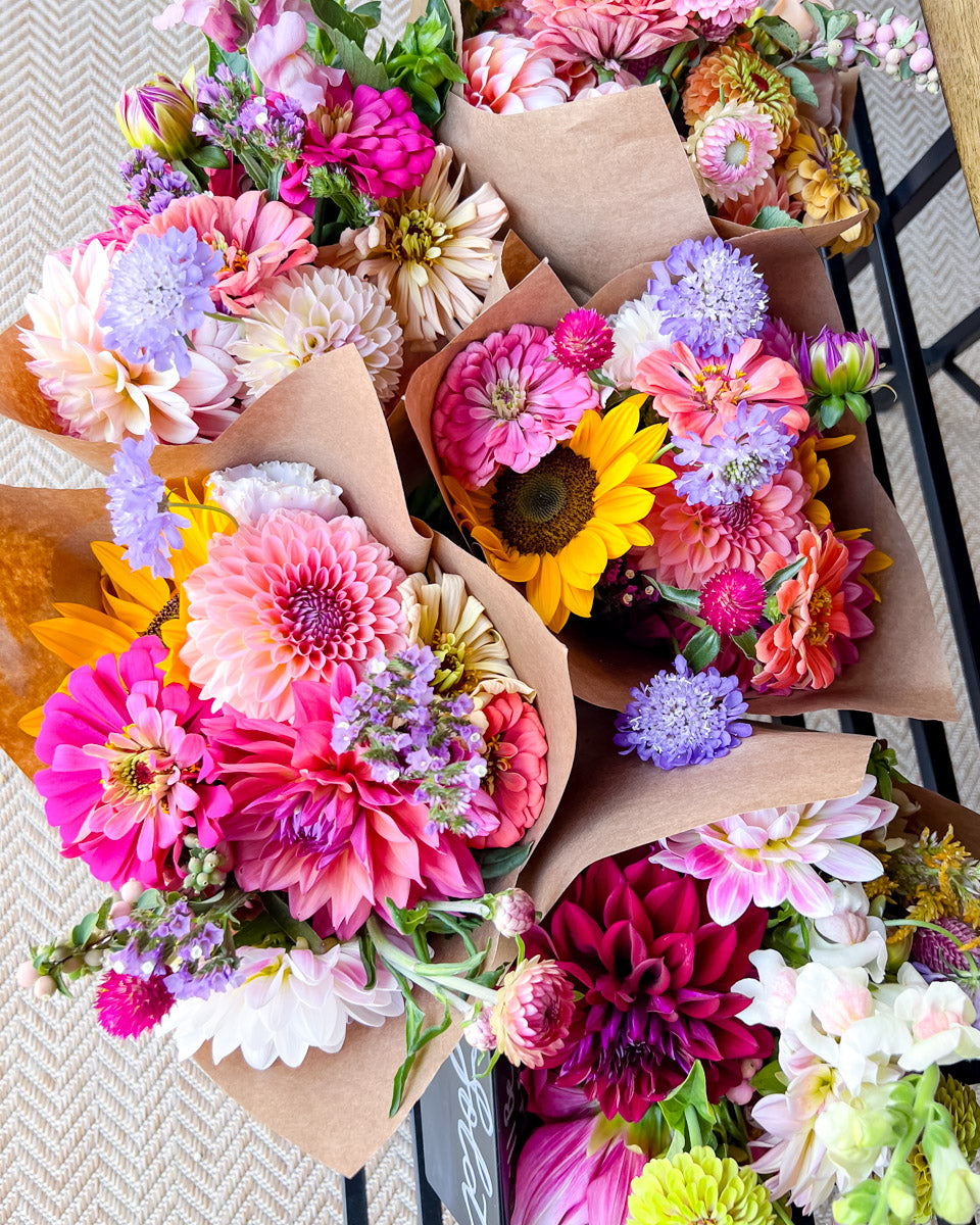 A close-up overhead view of several small flower bouquets wrapped in brown kraft paper, filled with colorful blooms including pink, purple, yellow, white, and peach flowers, arranged on a black shelf over a textured beige surface.