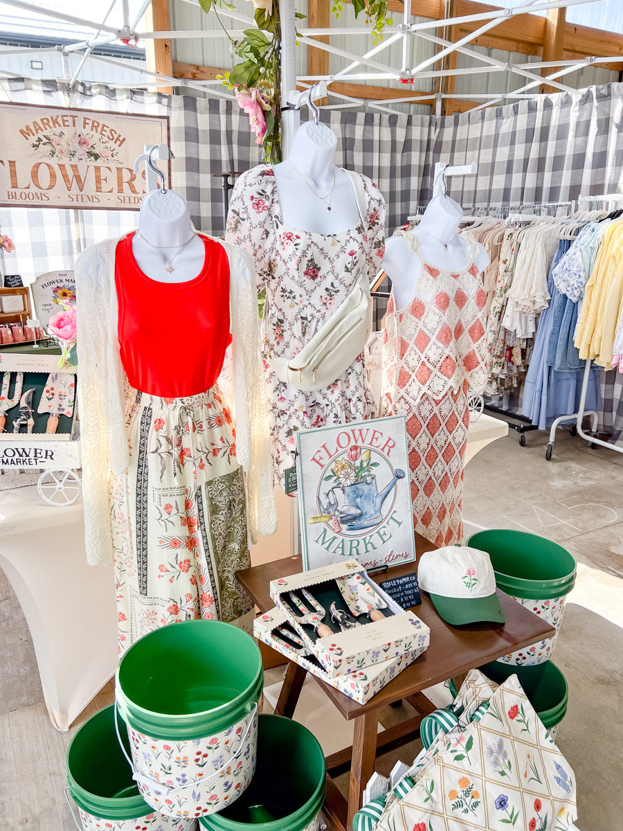 A boutique display featuring three mannequins dressed in vintage-style floral and patterned dresses, alongside matching patterned planters, a small table with floral accessories, and a checkered market canopy backdrop.