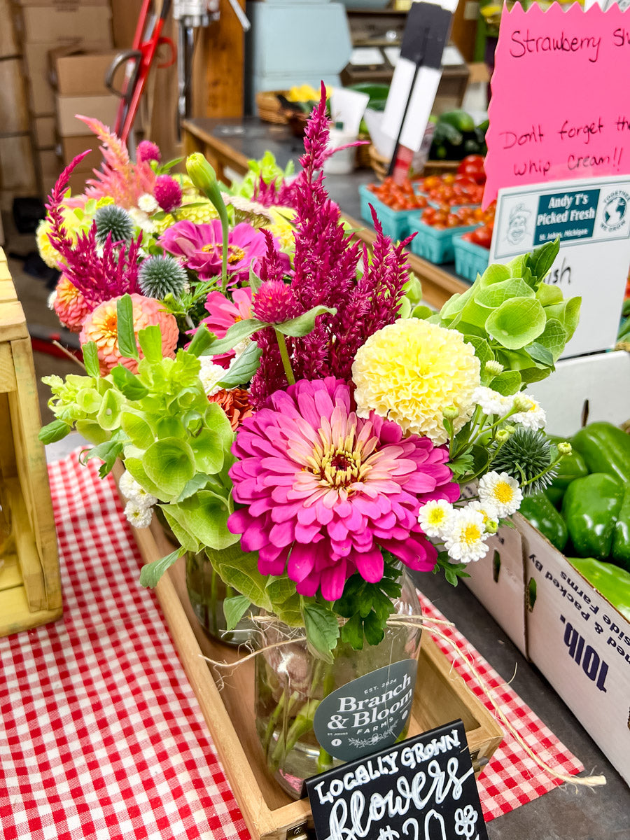 A vibrant, colorful flower bouquet in a clear glass jar with a wooden crate base, displayed on a red and white checkered tablecloth at a market. The bouquet features bright pink, yellow, green, and purple flowers, with a handwritten sign in front reading 'Locally Grown Flowers $20.