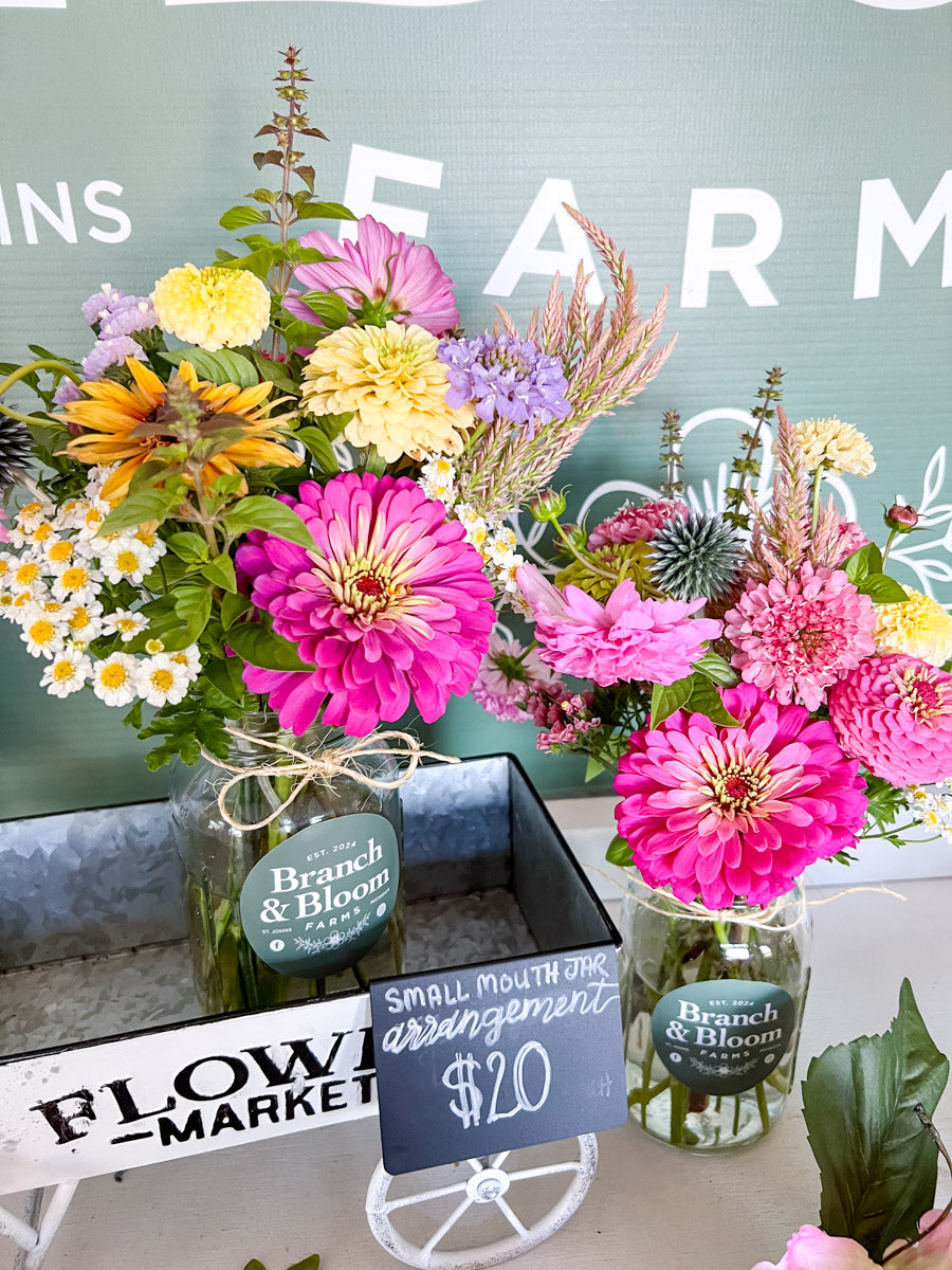 Two colorful floral arrangements in glass jars displayed on a table, with a small chalk sign reading $20 and a rustic sign in the foreground. The background features a painted green wall with white lettering.