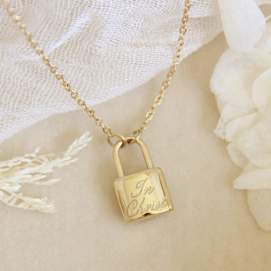 Gold lock-shaped pendant necklace with 'In Christ' engraving on a soft white background