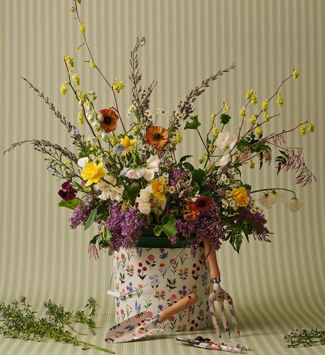 Floral arrangement in a floral printed plastic bucket with gardening tools on a striped background