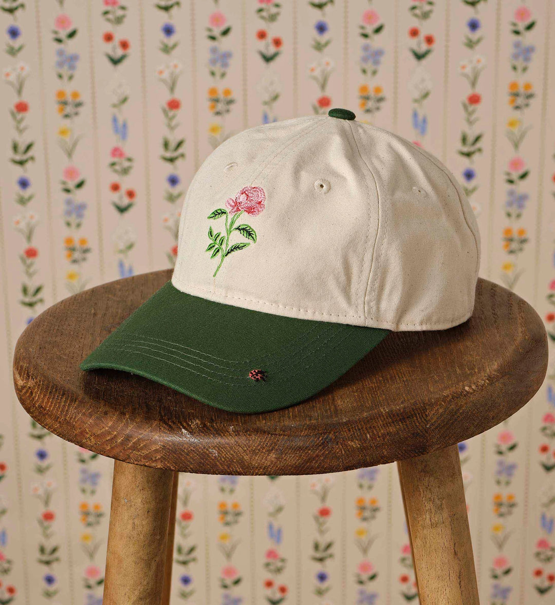 Two-tone cap with floral embroidery on a wooden stool against a floral-patterned wall.