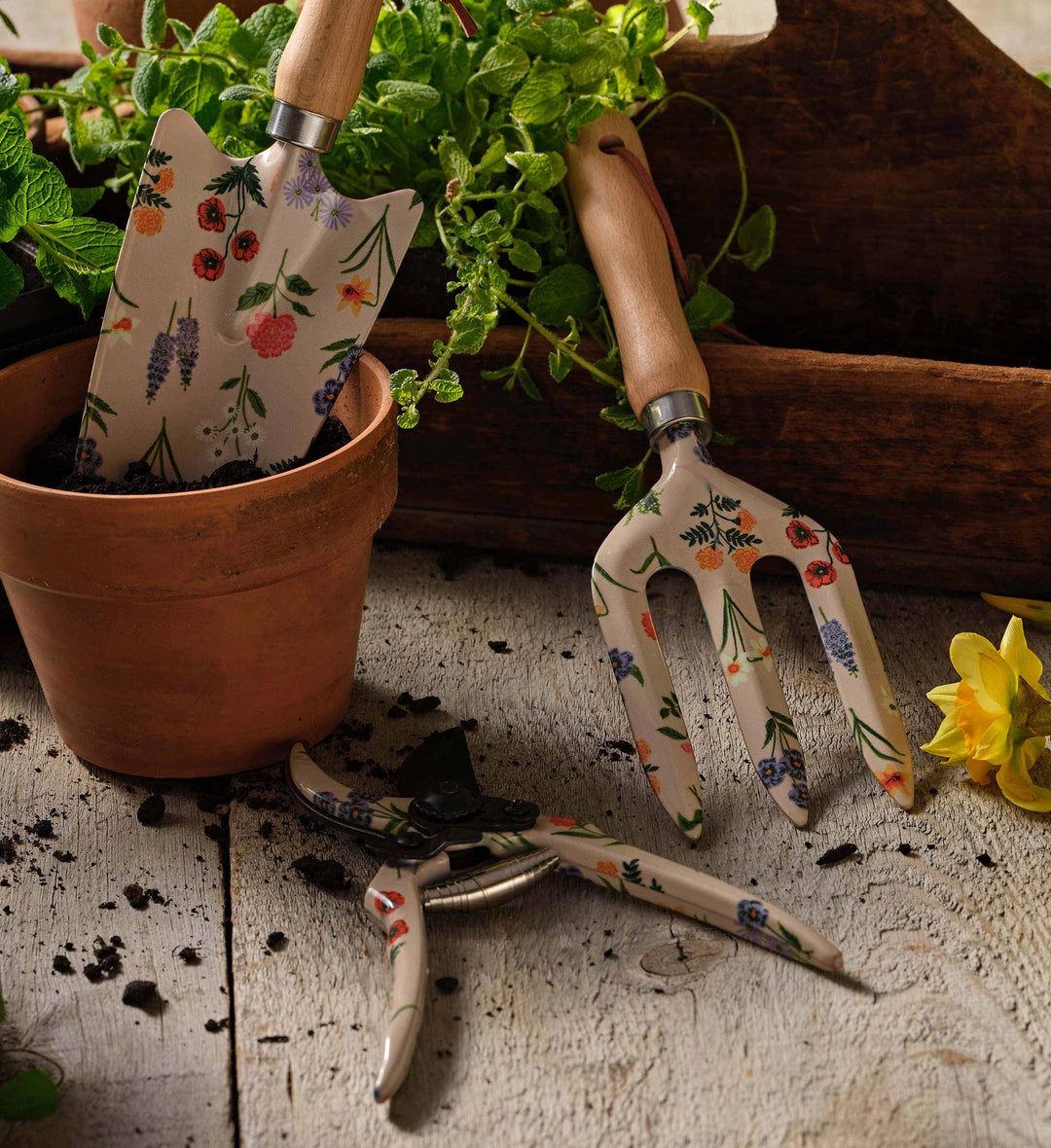Gardening tools with floral handles on a wooden surface with plants and a pot.